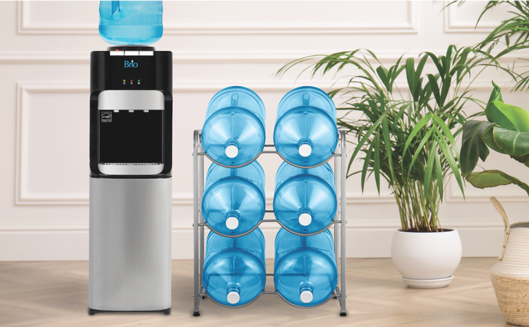Brio water dispenser with a blue water jug on top, next to a metal rack holding six blue water jugs, with potted green plants in the background.